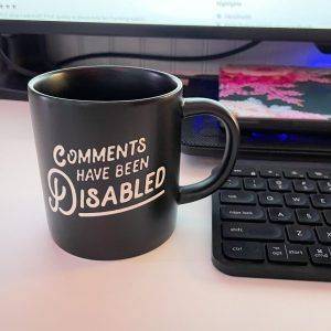 Eine Tasse mit der Aufschrift “Comments Have Been Disabled”, daneben eine Tastatur und dahinter ein Laptop auf einer Kühlunterlage.